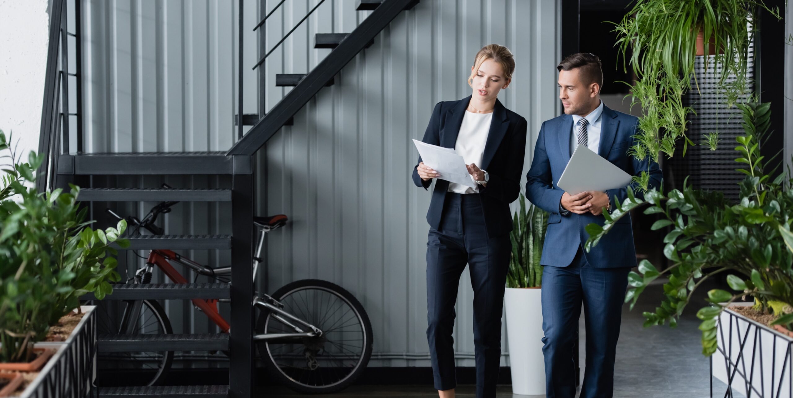 Two business professionals walk together through an office with modern decor. The woman holds documents, and the man, a government contracting recruiter, carries a laptop. They are engaged in discussion as they walk past a staircase and greenery, with a bicycle visible in the background.
