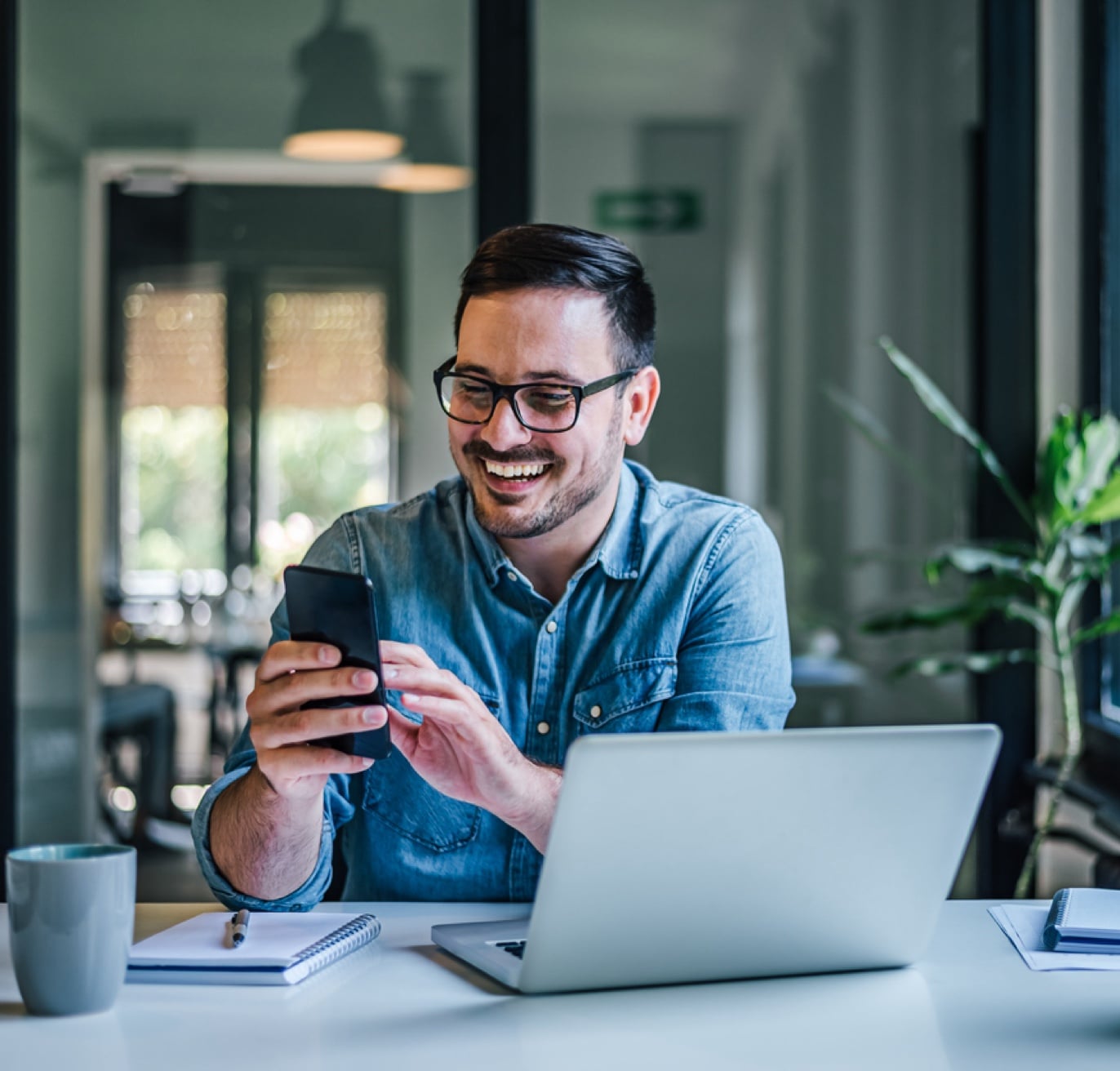 A man wearing glasses and a denim shirt is sitting at a desk, smiling while holding a smartphone. In front of him, there's a laptop, a notebook, and a mug. In the background, there's a glass wall and a potted plant. He appears to be engrossed in government contracting executive search activities.