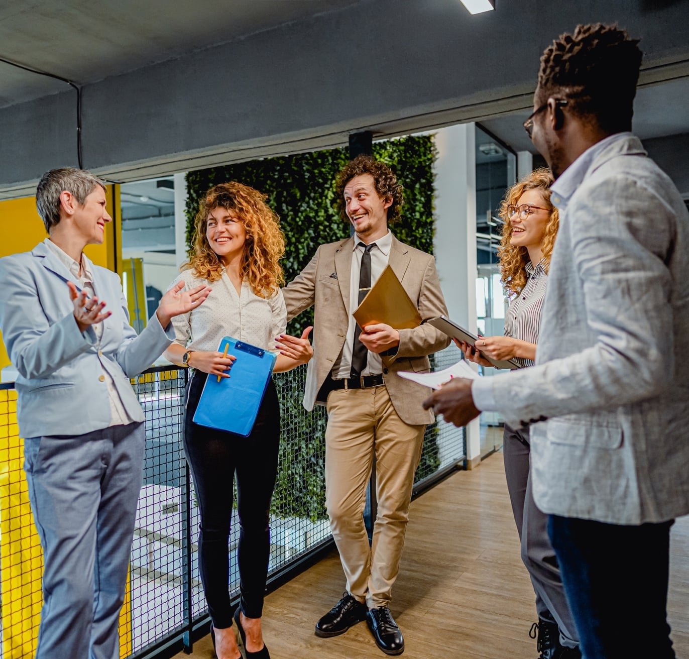 A diverse group of five professionals are engaged in a lively discussion in a modern office corridor. They are smiling and holding documents, possibly related to government contracting. A green wall with plants is in the background, adding a natural element to the contemporary setting.