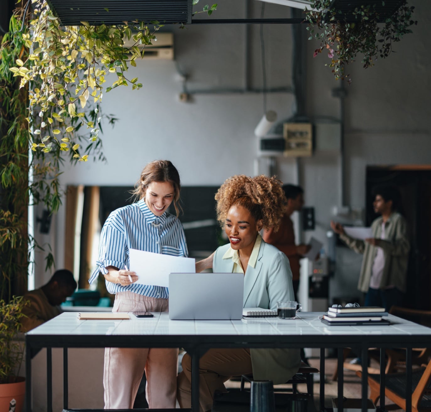 Two women are collaborating at a desk in an open, plant-filled workspace. One is standing and holding papers, smiling, while the other is seated with a laptop, also smiling. The vibrant atmosphere buzzes with activity as the team focuses on technology recruiting efforts in the background.