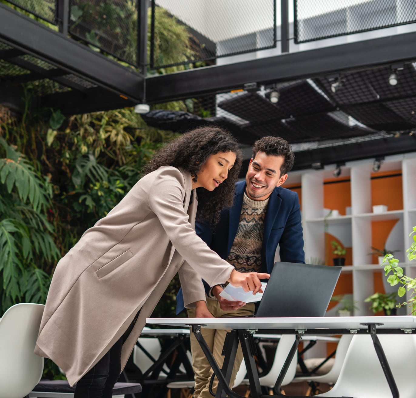 Two colleagues collaborate intently while looking at a laptop screen in a modern office with a green, plant-filled background. One points at the screen while the other, possibly a tech recruiter, smiles, suggesting a positive and engaging discussion.