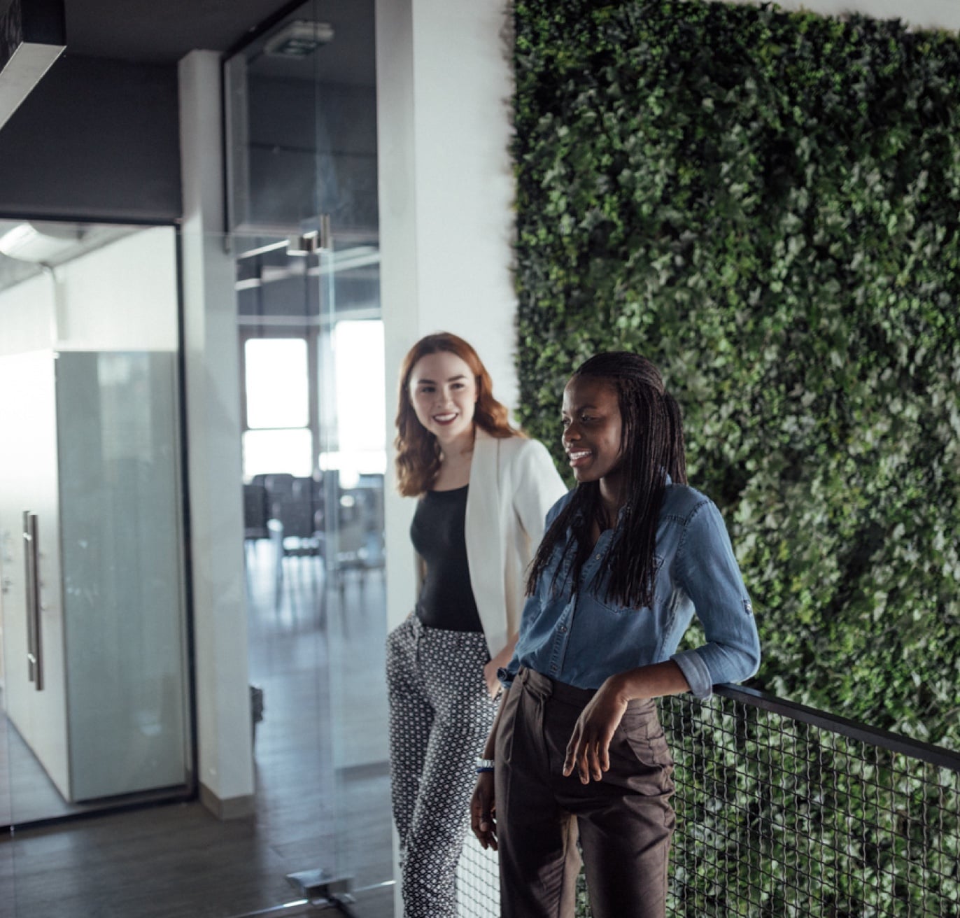 Two women are walking and conversing in an office corridor. One is wearing a white blazer and black and white polka dot pants, and the other is dressed in a blue shirt and brown pants. They are passing by a vertical garden wall inside the modern, well-lit technology recruiting office space.