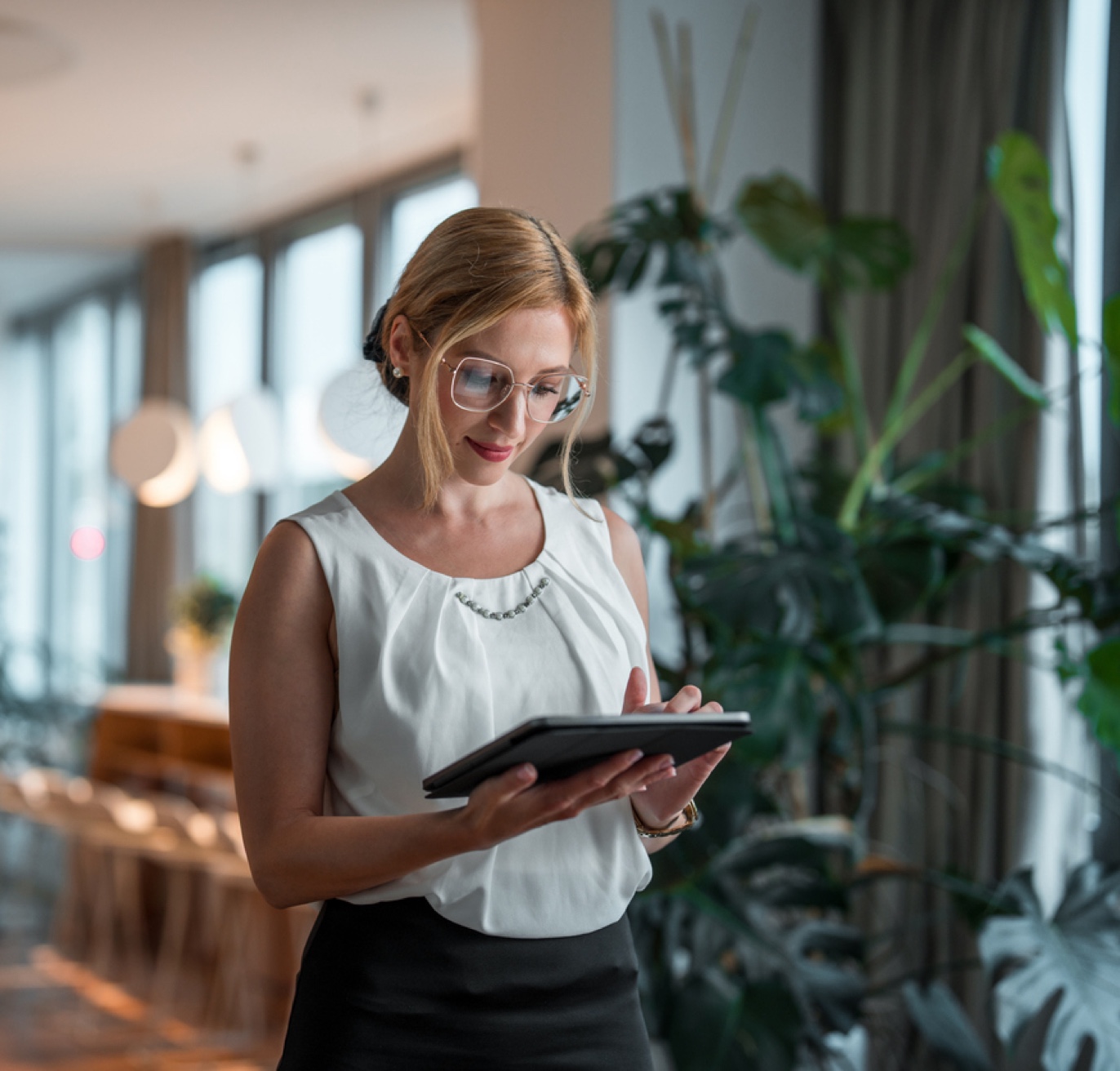 A businesswoman with glasses and a sleeveless white blouse uses a tablet in a modern office with large windows and lush green plants. She appears focused as she navigates government contracting opportunities, the atmosphere well-lit with a natural and professional ambiance.