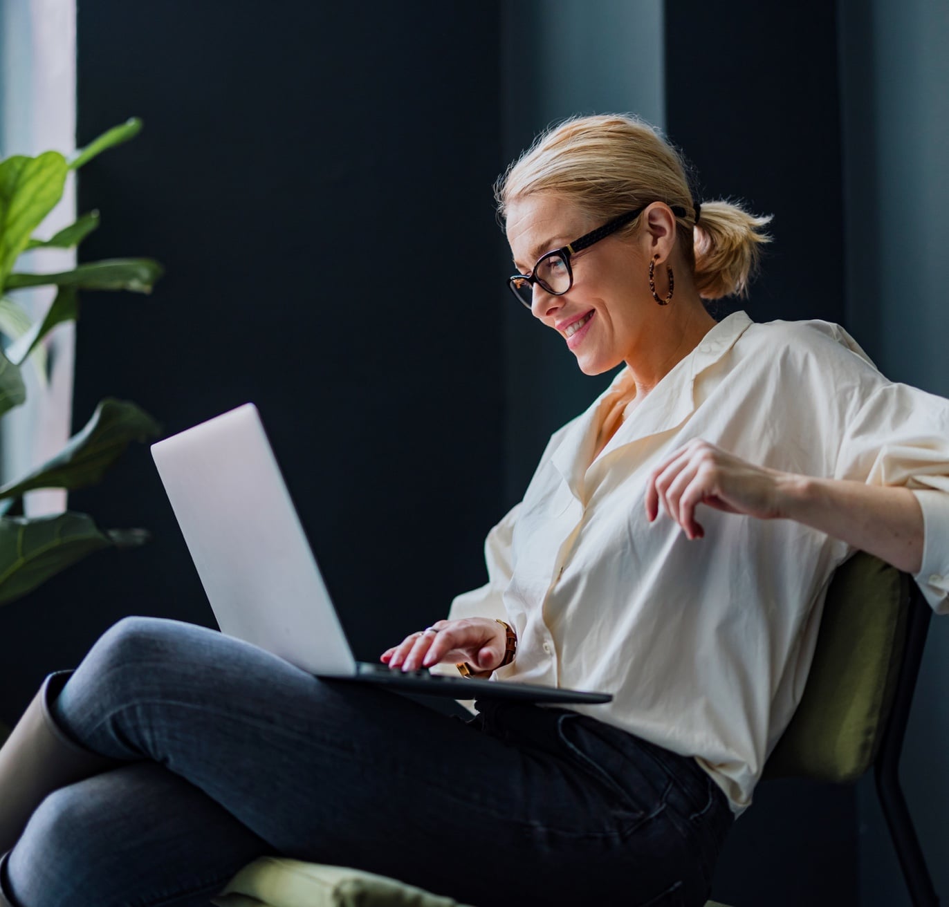 A tech recruiter with blonde hair tied back, wearing glasses, a white blouse, and dark jeans, is sitting on a chair in front of a laptop. She is smiling and appears to be engrossed in what she is viewing. There is a green plant in the background.